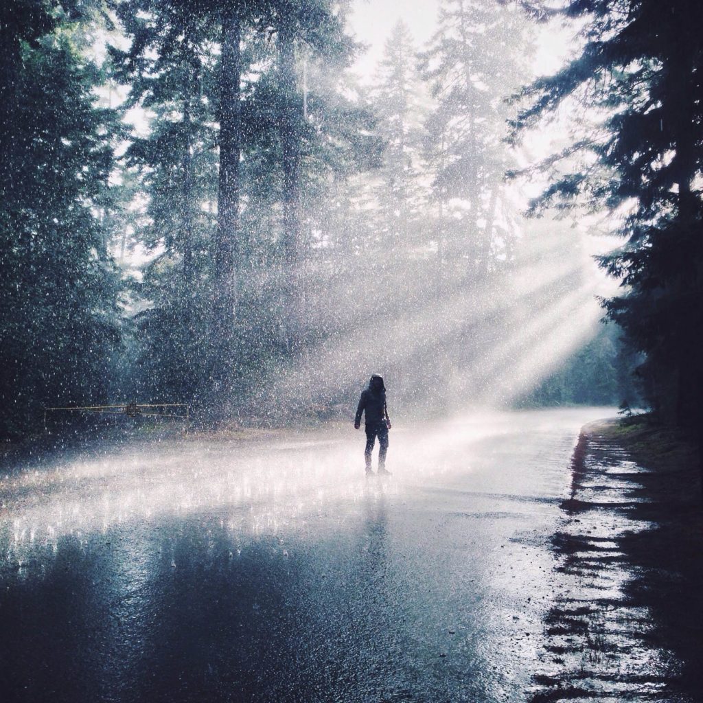 A man stands in the middle of a road as the rain is caught by rays of sunlight.
