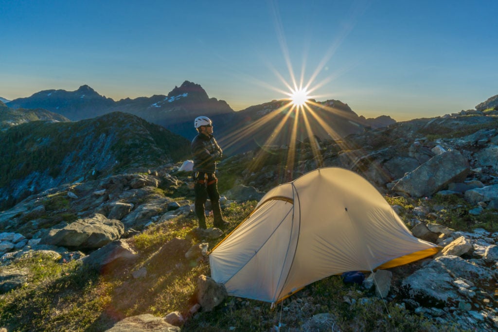 A man wearing a white helmet stands next to a tent set up in a rocky landscape at sunset.