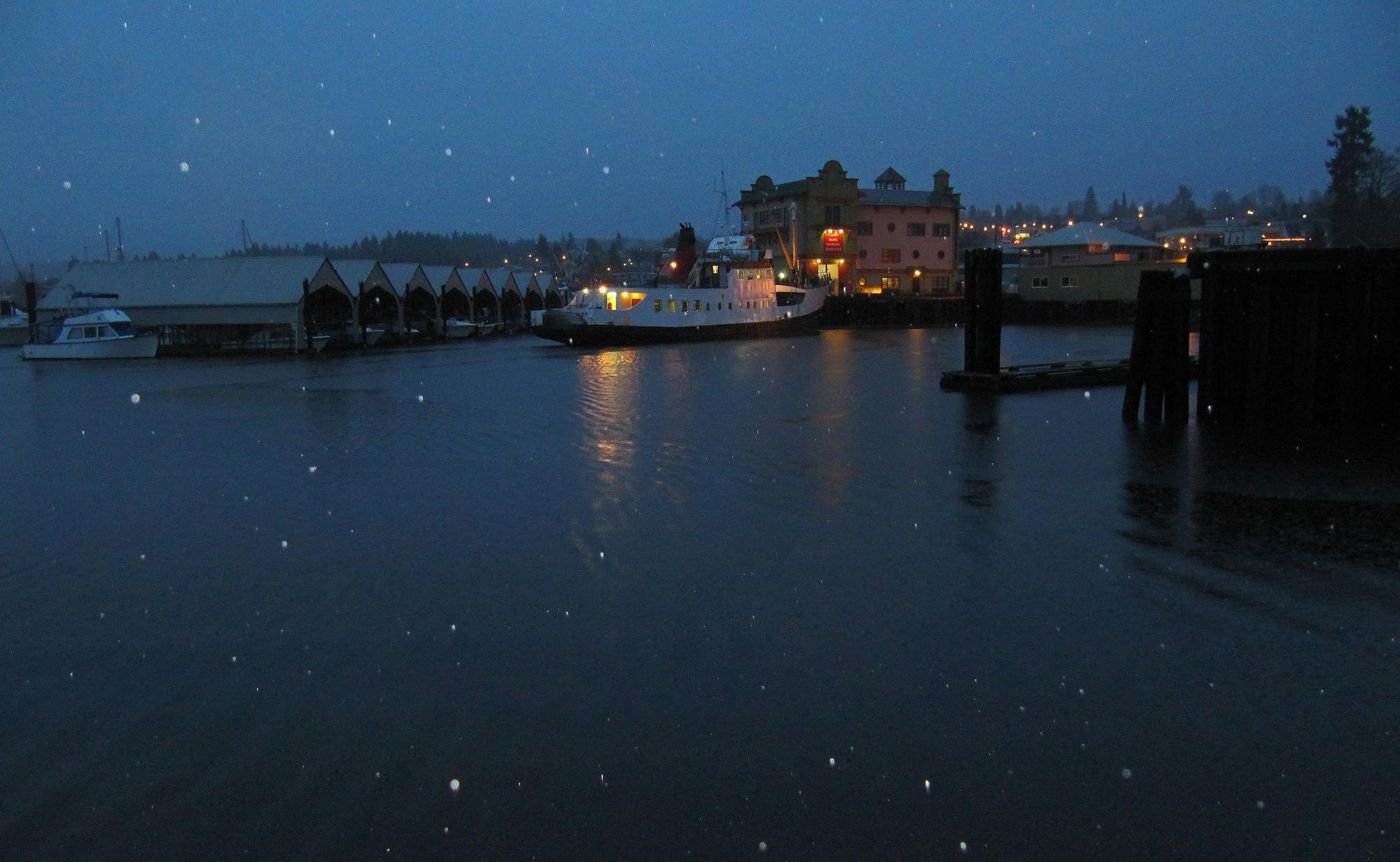 Snow softly falls over a quiet harbour at night.