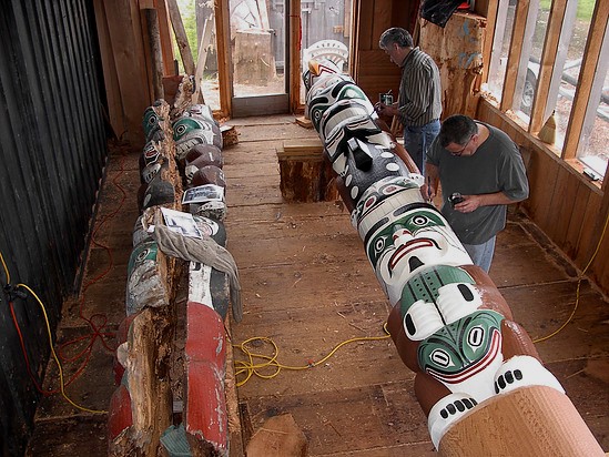 Two men paint a totem pole in green, white, brown and black.