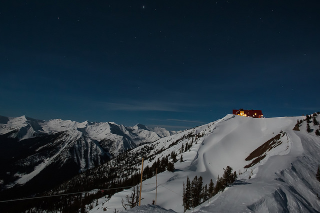 A brightly lit cabin sits at the top of a snow-covered mountain.