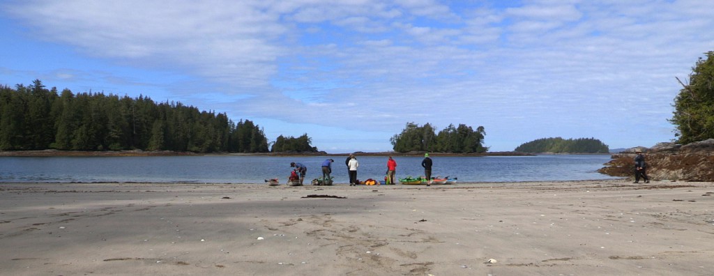 A group of kayakers stand on a beach at the water’s edge.