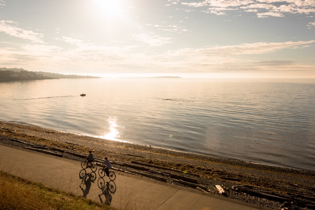 Two cyclist pedal on a path along the ocean at sunset.
