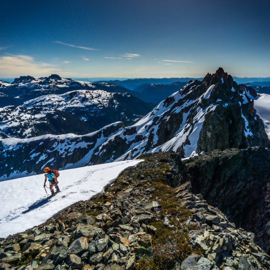 A hiker traverses a snow-covered rocky landscape with snow-capped mountains in the background.