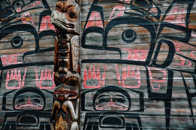 An intricately carved totem pole stands in front of a colourfully painted wooden wall.