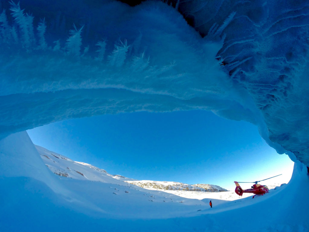 A red helicopter touches down in a snowy landscape, in front of an ice cave.