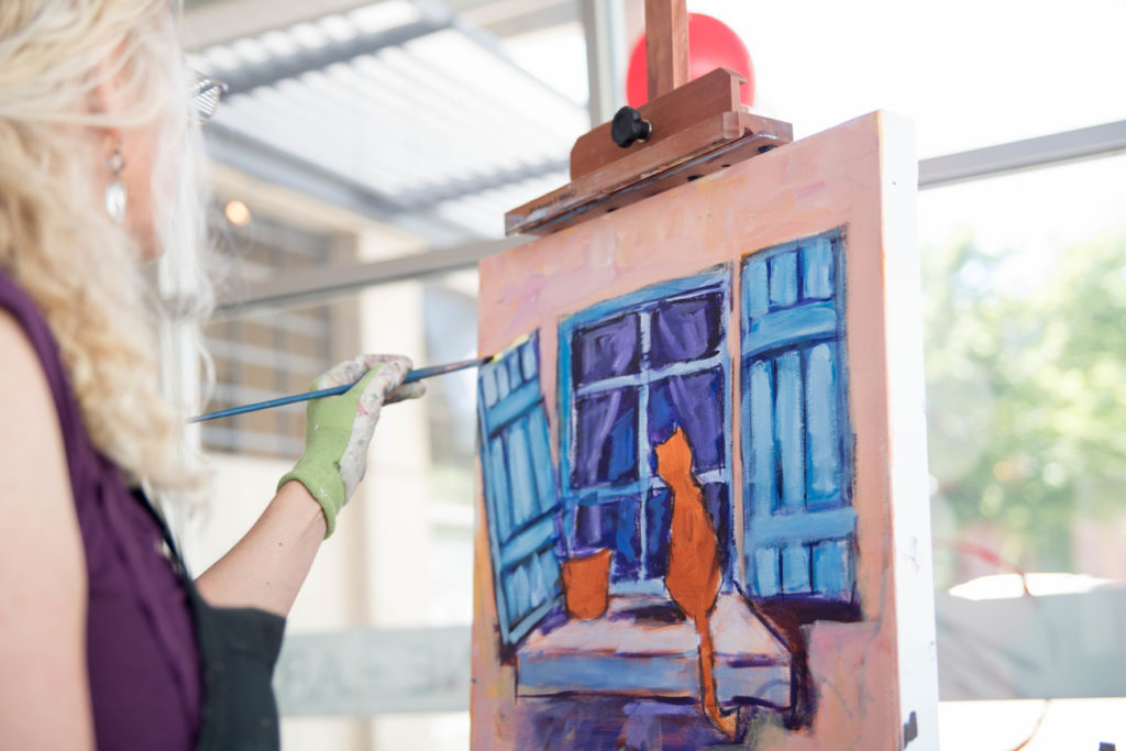 An artist at her easel, painting an orange cat looking out the window.