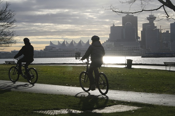 Two cyclists cycling along the Stanley Park Seawll in Vancouver on a cloudy day with the city skyline in the background.