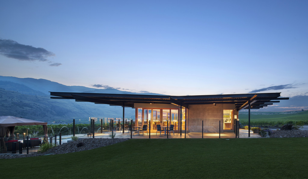 Black Hills Estate Winery lit up at dusk, with a flat modern roof, chairs outside on a patio, the Okanagan hills in the background and a lush green lawn in the foreground.