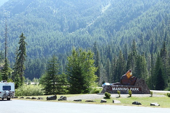 The entrance to Manning Provincial Park, with a wooden sign, an RV driving down the road and thick forested hills in the background.