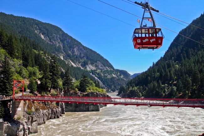 A red airtram travels over a red bridge and rushing waters.