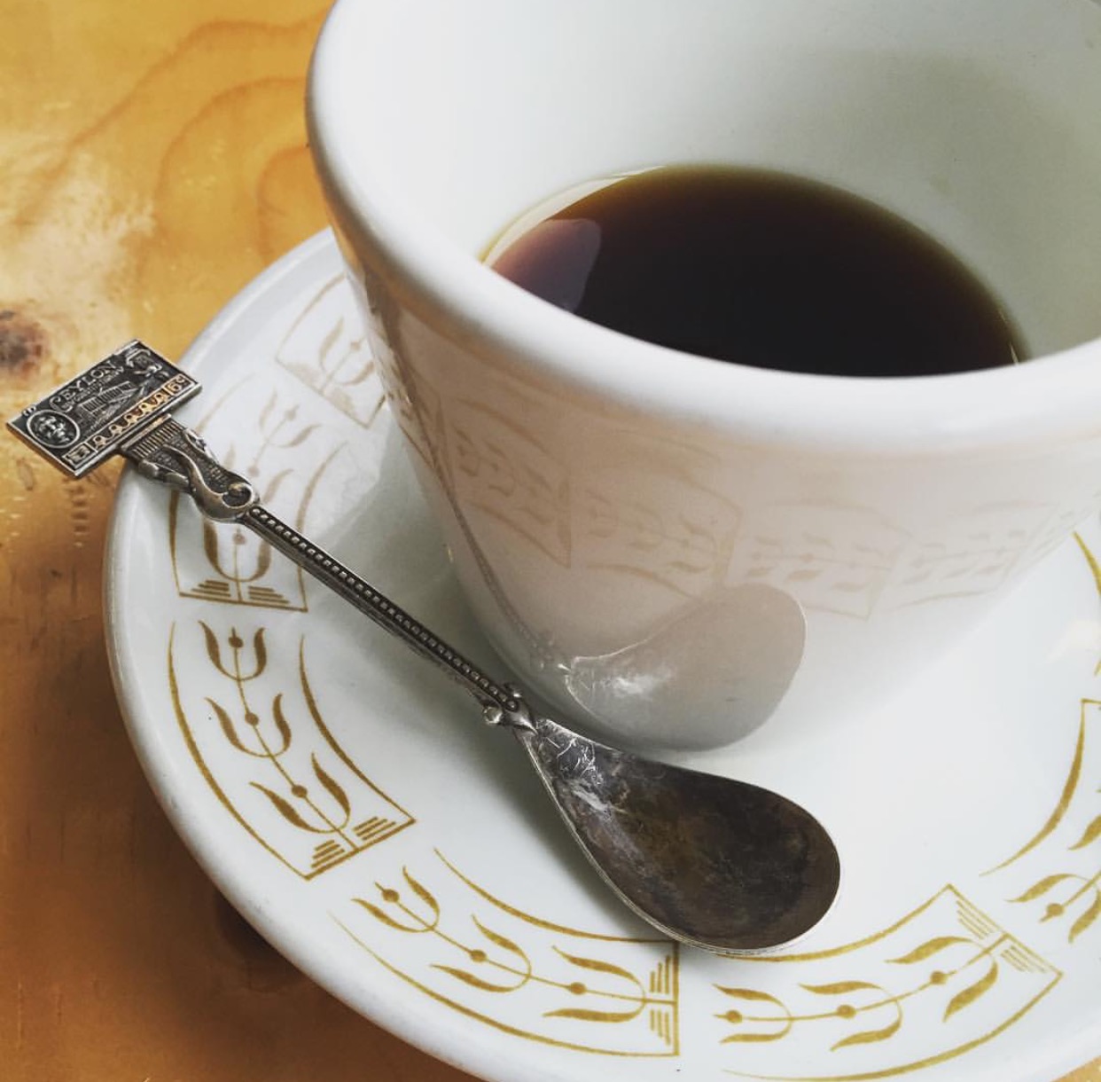 A cup of coffee on a white and gold saucer with a small spoon.