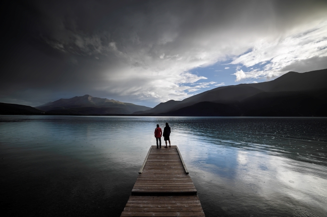 Two people stand at the edge of a dock, taking in the mountain views as evening creeps in.