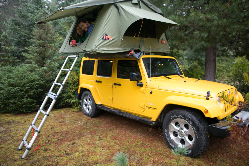 A man and woman lay in a fold-out roof-tent on top of a yellow Jeep