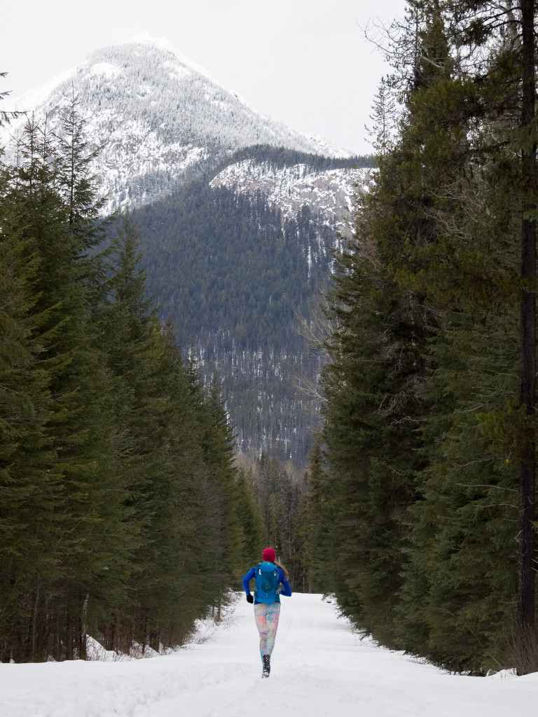 A woman wearing a backpack runs down a snow-covered trail lined with pine trees.