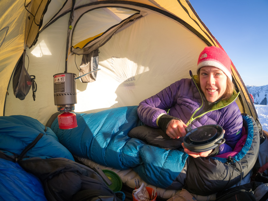 A woman in a blue sleeping bag wears a winter jacket and toque inside her tent.