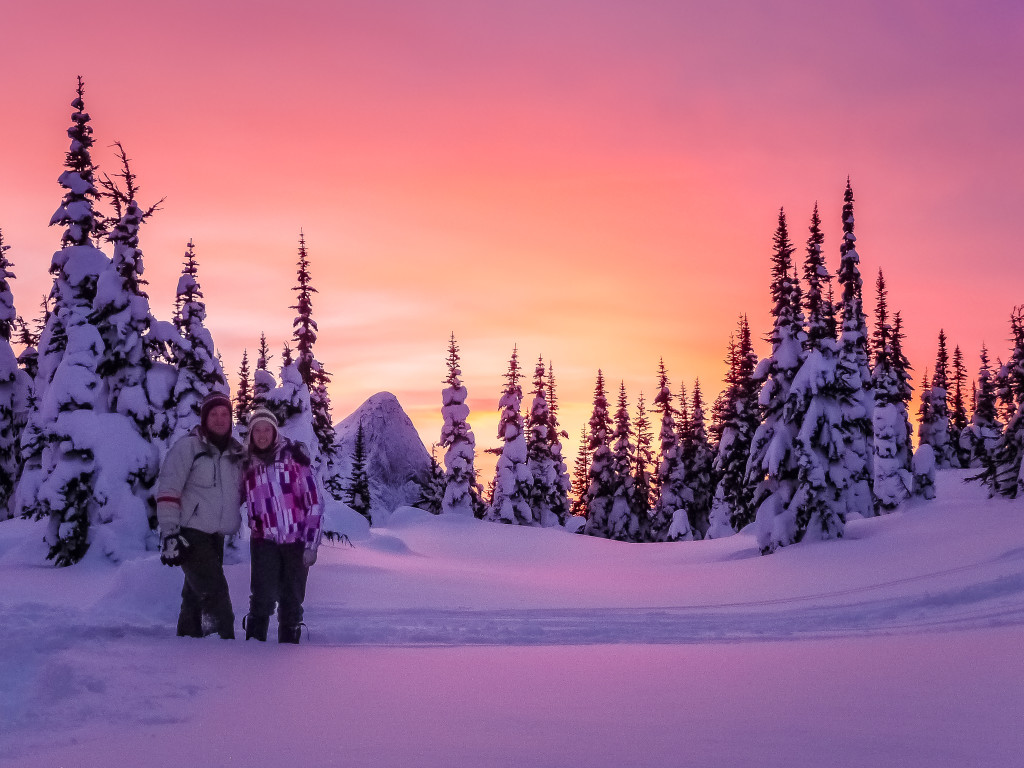 A man and a woman stand in a snow-covered landscape under a stunning pink and orange sunset.
