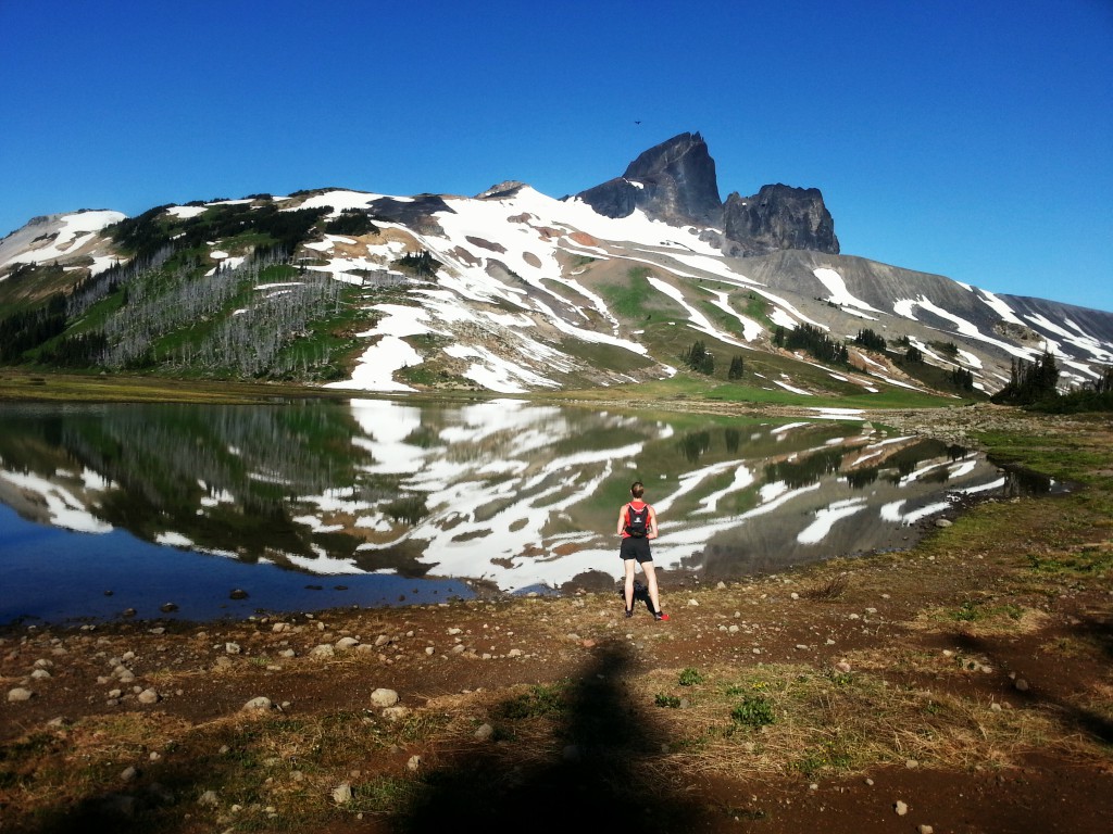 A hiker pauses to take in the reflection of a snow covered mountain in a still pond.