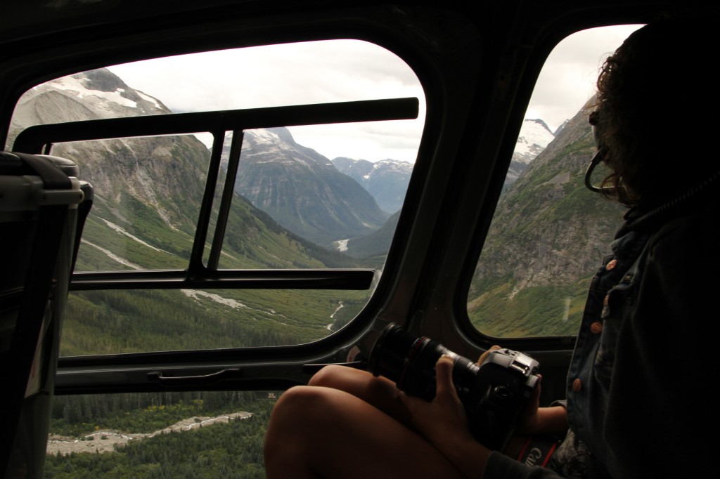 A woman sits in a helicopter, traveling over a mountain valley.