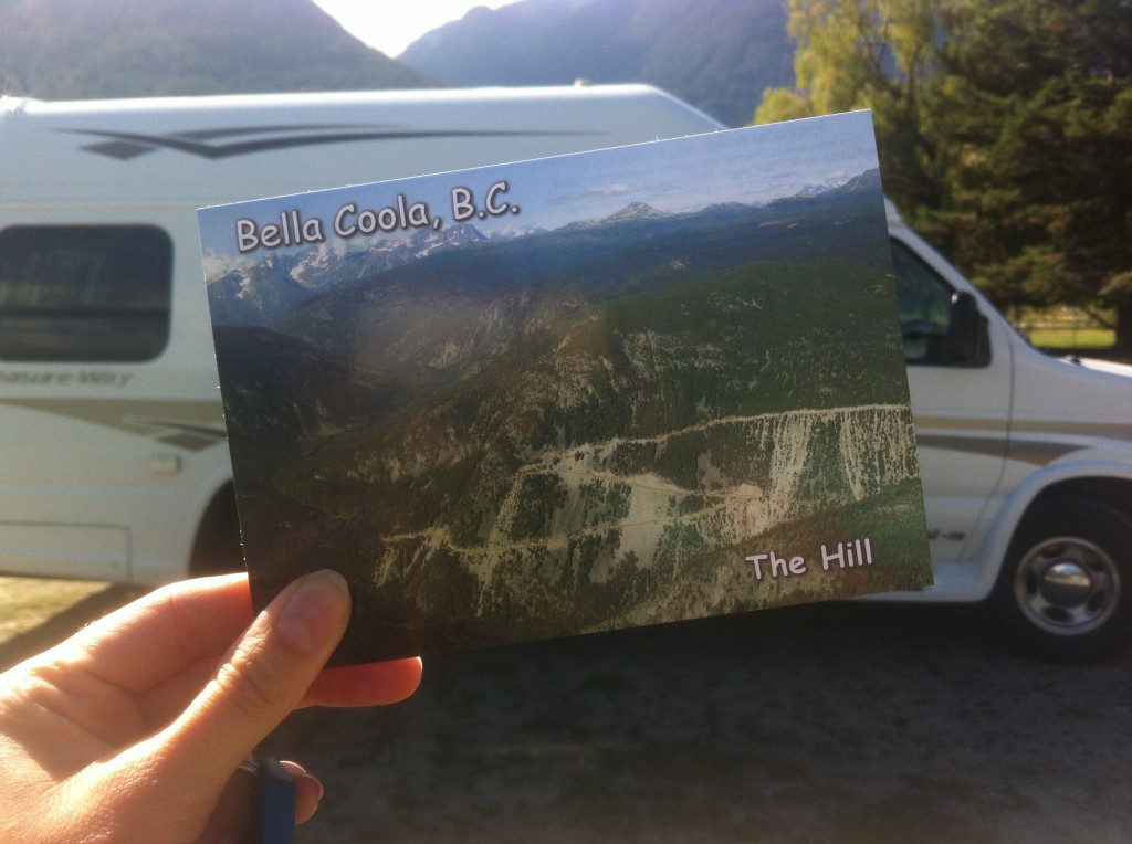 A hand hold up a postcard of a rolling landscape that says “Bella Coola, B.C. The Hill”.