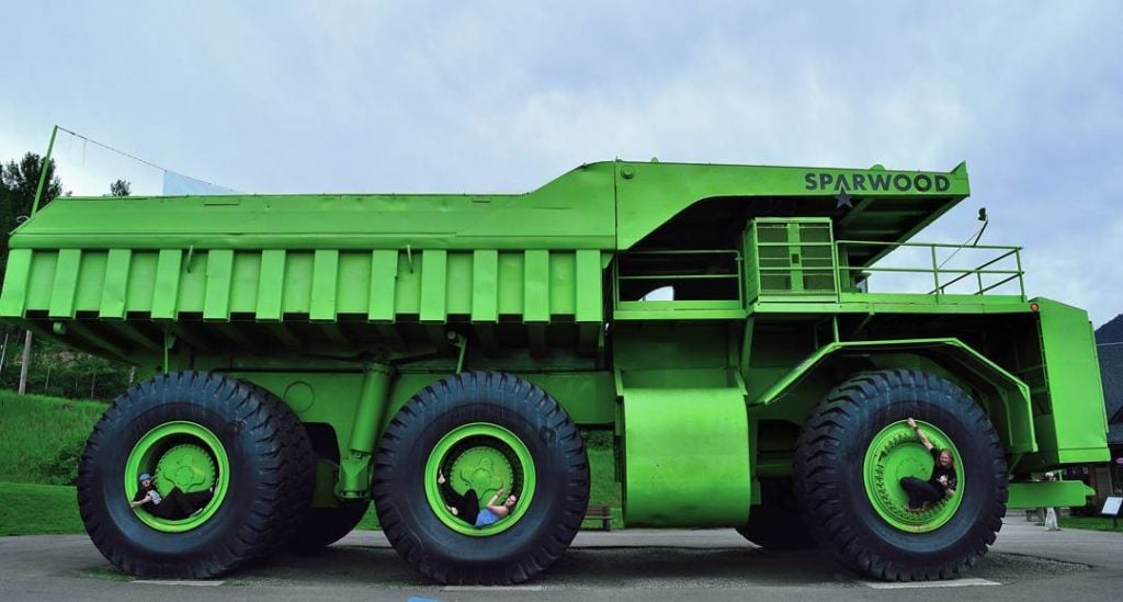Three people sit in the wheel spokes of a giant green truck.
