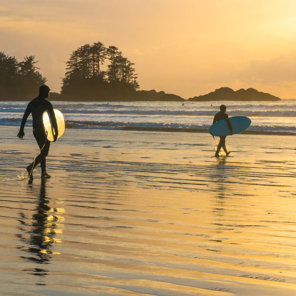 Two surfers carry their boards to the water under a golden sunrise.