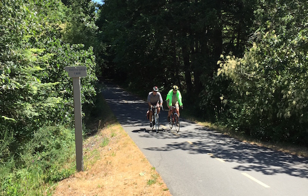 Two cyclists cycling towards the camera, one wearing a bright green jacket, as they head to the turn-off for Thetis Lake on the Galloping Goose Regional Trail.