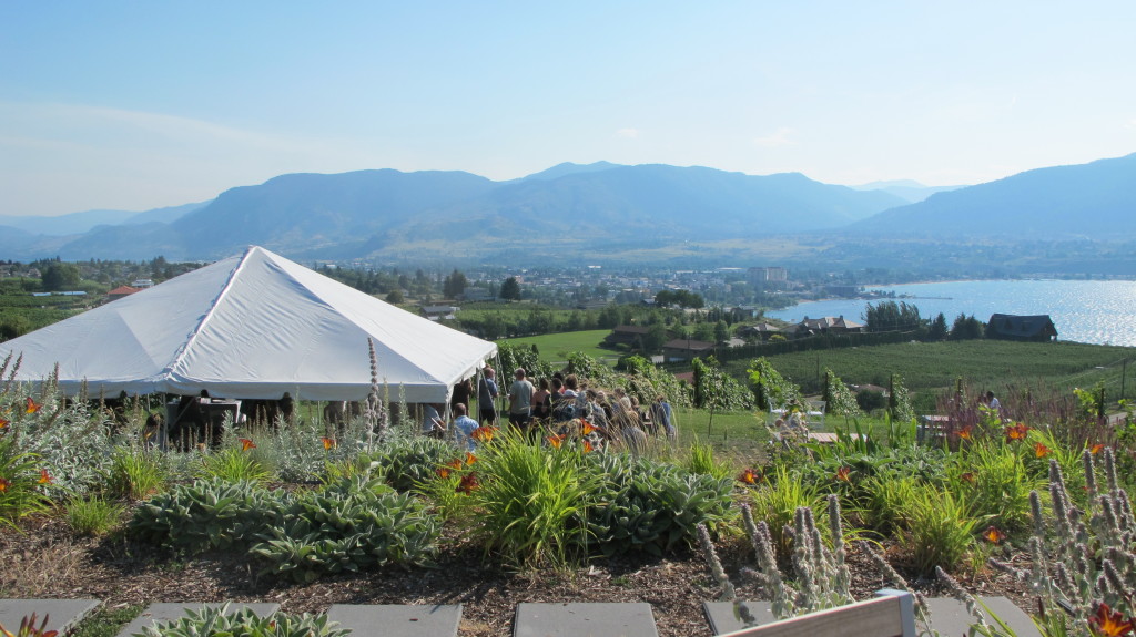 A white tent and a group of people stand on a lush lawn with views of the water and mountains.