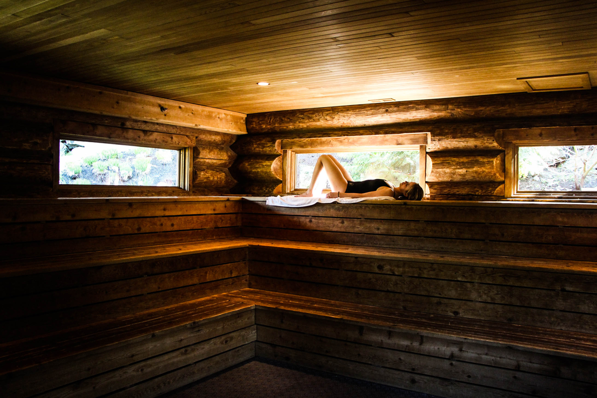 A woman lies down on a towel inside a dry sauna at the Scandinave Spa in Whistler. She is wearing a black one piece bathing suite and is alone in the sauna.
