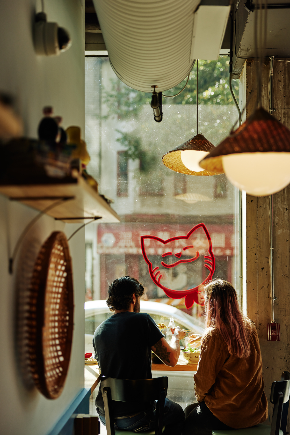 Two people sit together facing a window at Fat Mao Noodles in Chinatown. They are sitting on barstools at a window ledge and a red cat is outlined on the window.