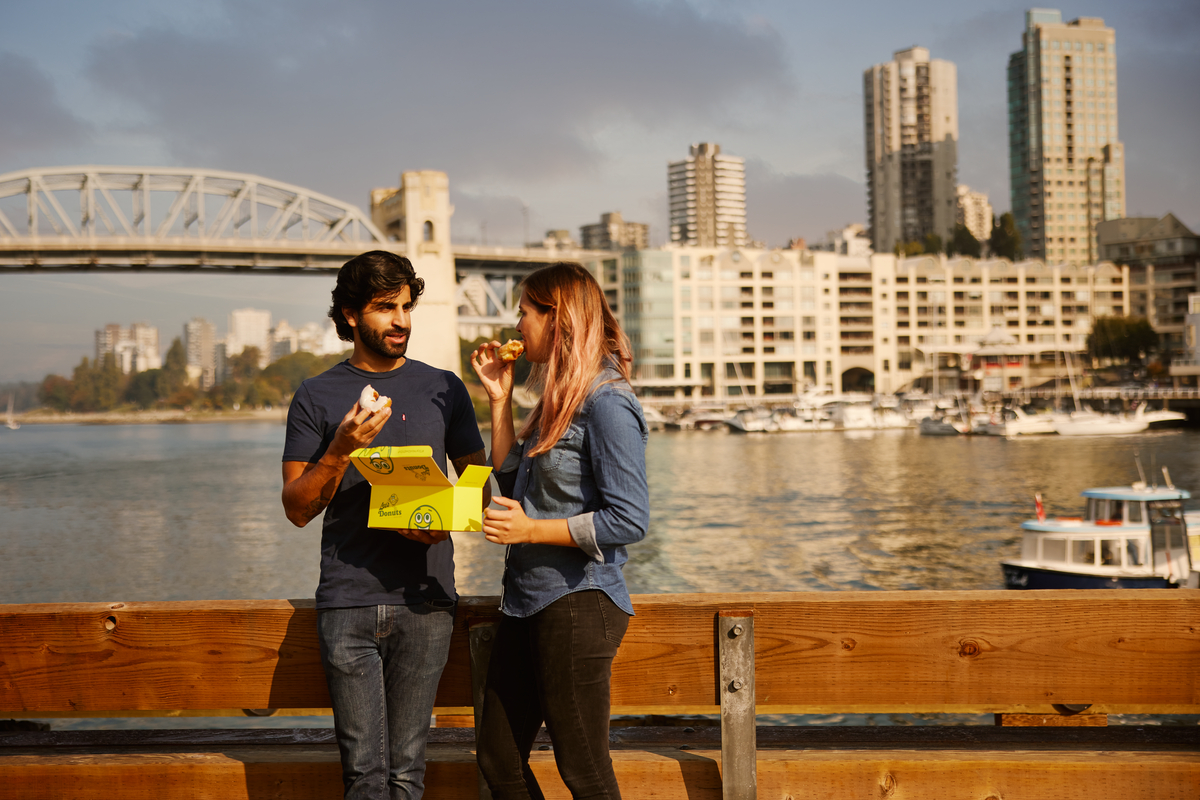 Two people Enjoying Lee's Donuts on the water's edge at Granville Island Public Market. The Burrard Bridge and and skyscrapers are seen in the background.