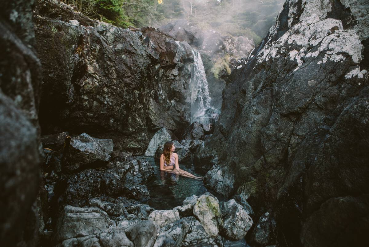 A woman sits in a natural hot spring near Tofino. A small waterfall is behind her.