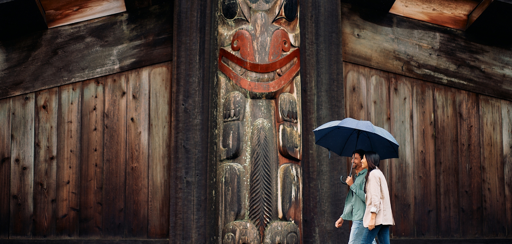 Two people walk under a blue umbrella with a weathered Indigenous pole and building behind them.