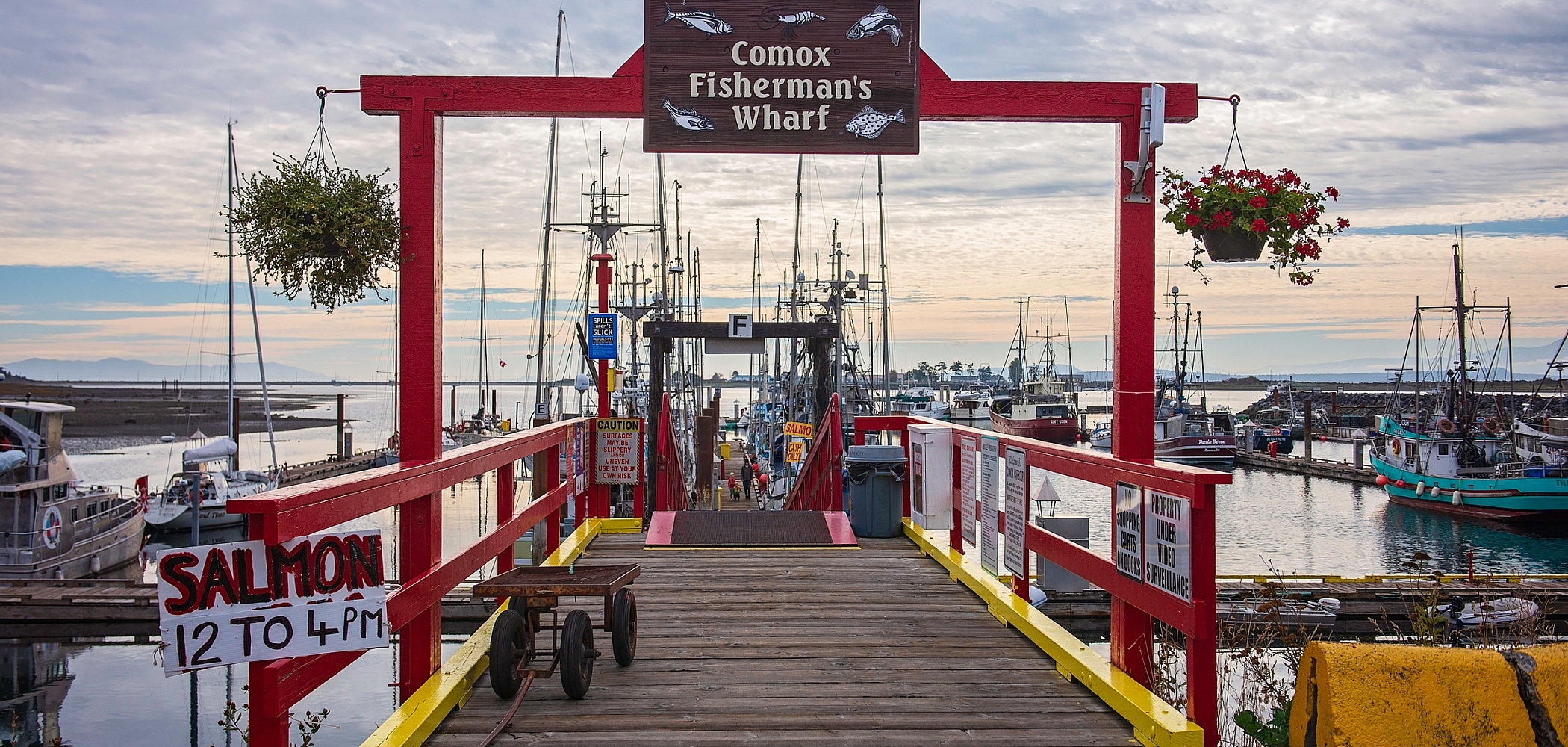 Brightly-coloured red and yellow archway and railing leading out to a dock lined with fishing vessels. A sign reads, "Comox Fisherman's Wharf."