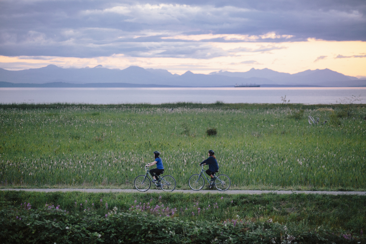 Two people cycle the West Dyke Trail as the sunsets. There is a grassy field behind them that leads to the ocean. 