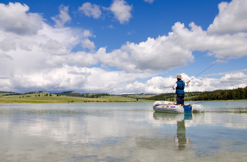 A man stands up in an inflatable dinghy, fly-fishing on a calm pond.