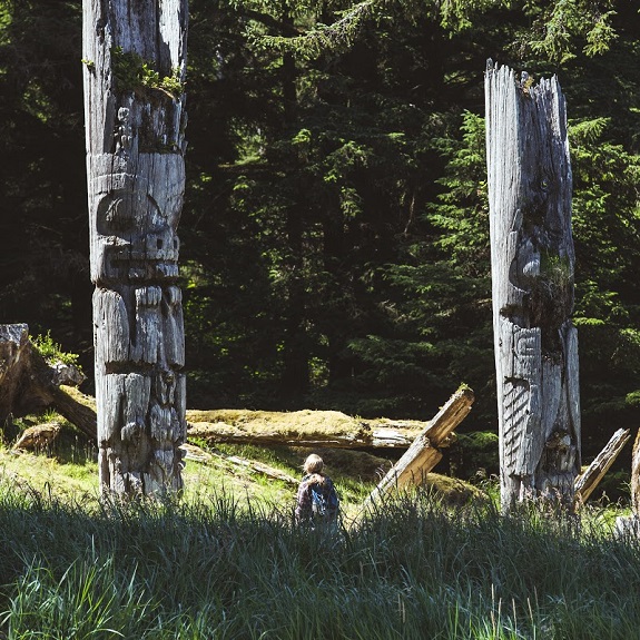 A hiker pauses to look at intricate carvings on two tall wooden poles.