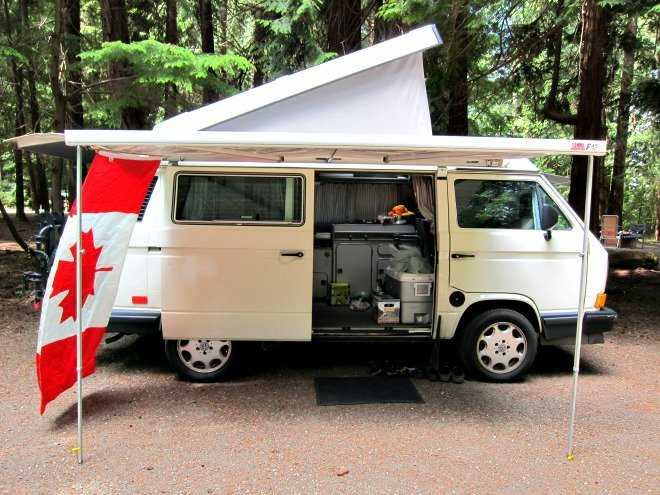 A beige camper van with a white awning and a Canadian flag.