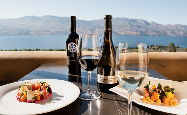 A table with a mountain view is set with two colourfully plated dishes, two bottles of wine, one glass of red wine, and one glass of white.
