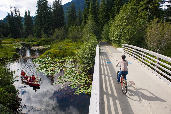 Two people in a canoe wave to a cyclist, pedaling across a bridge.