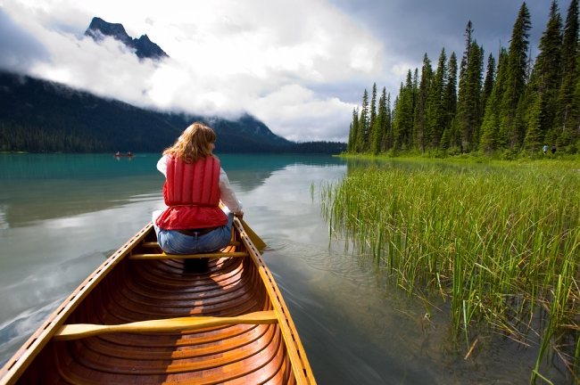 A woman paddles a canoe in calm waters, towards mist-covered mountains.