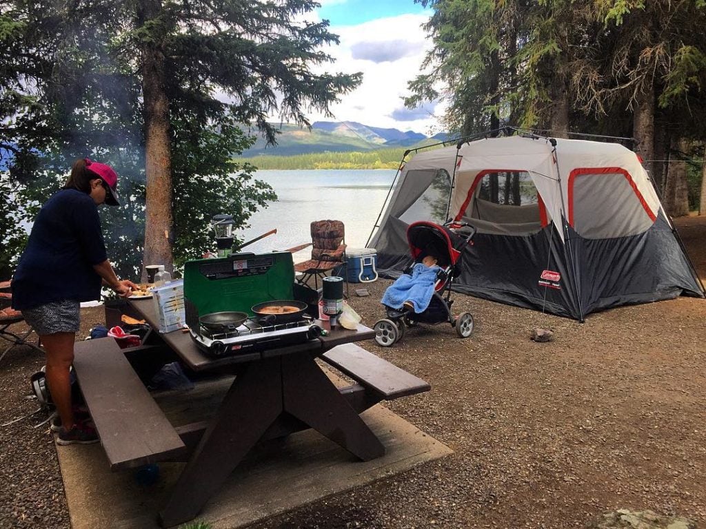 A woman prepares dinner at a campsite on the water with views of the mountains.