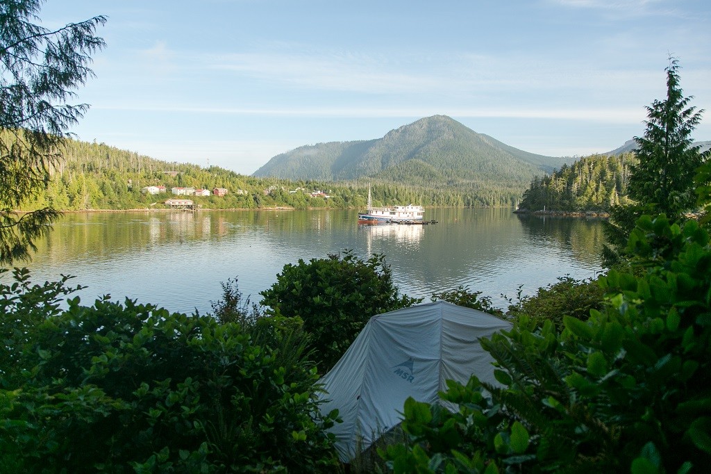 A tent is nestled within dense greenery overlooks a boat sailing in a small harbour.