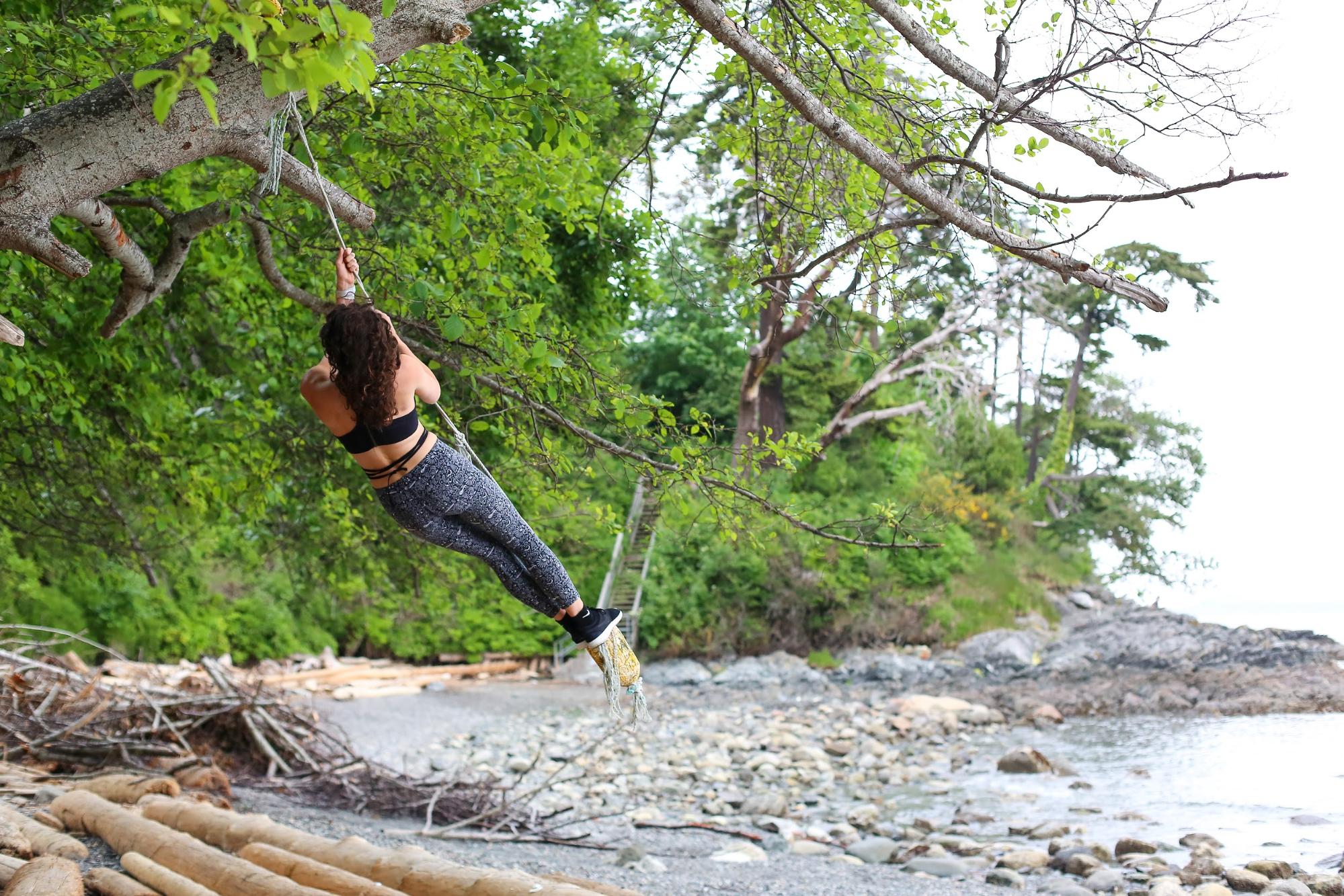 A woman swings from a rope tied to a tree over a rocky riverbank.