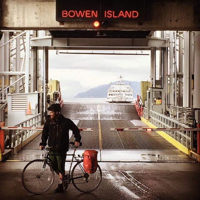 A man takes his bike across a ferry terminal with a sign in red that reads “Bowen Island”.