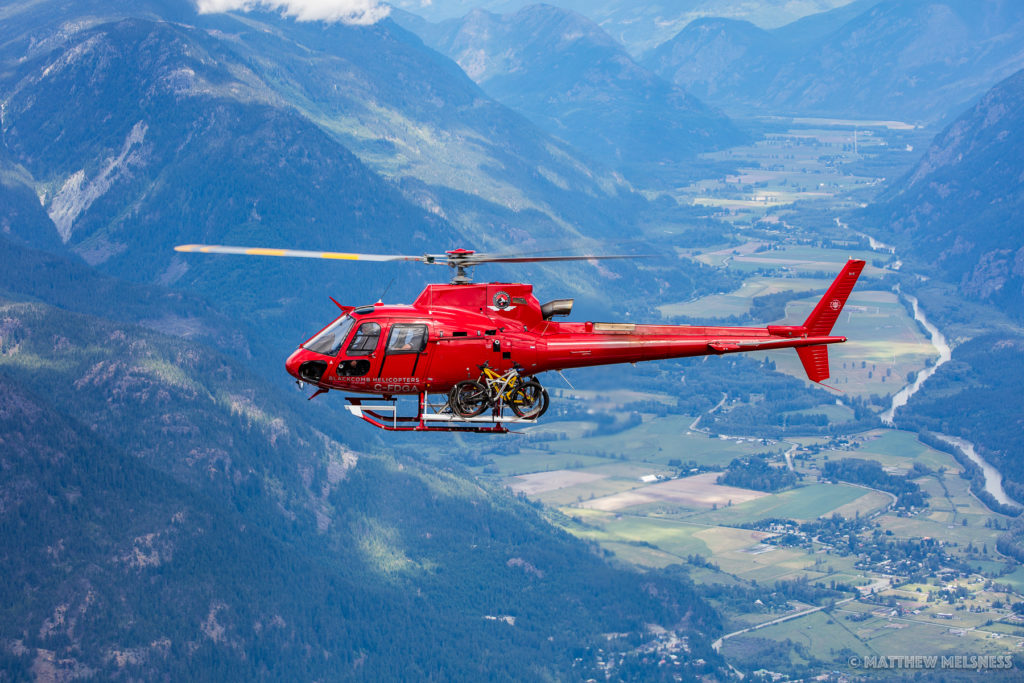 A red helicopter with mountain bikes strapped to the exterior flies over a mountain landscape. 