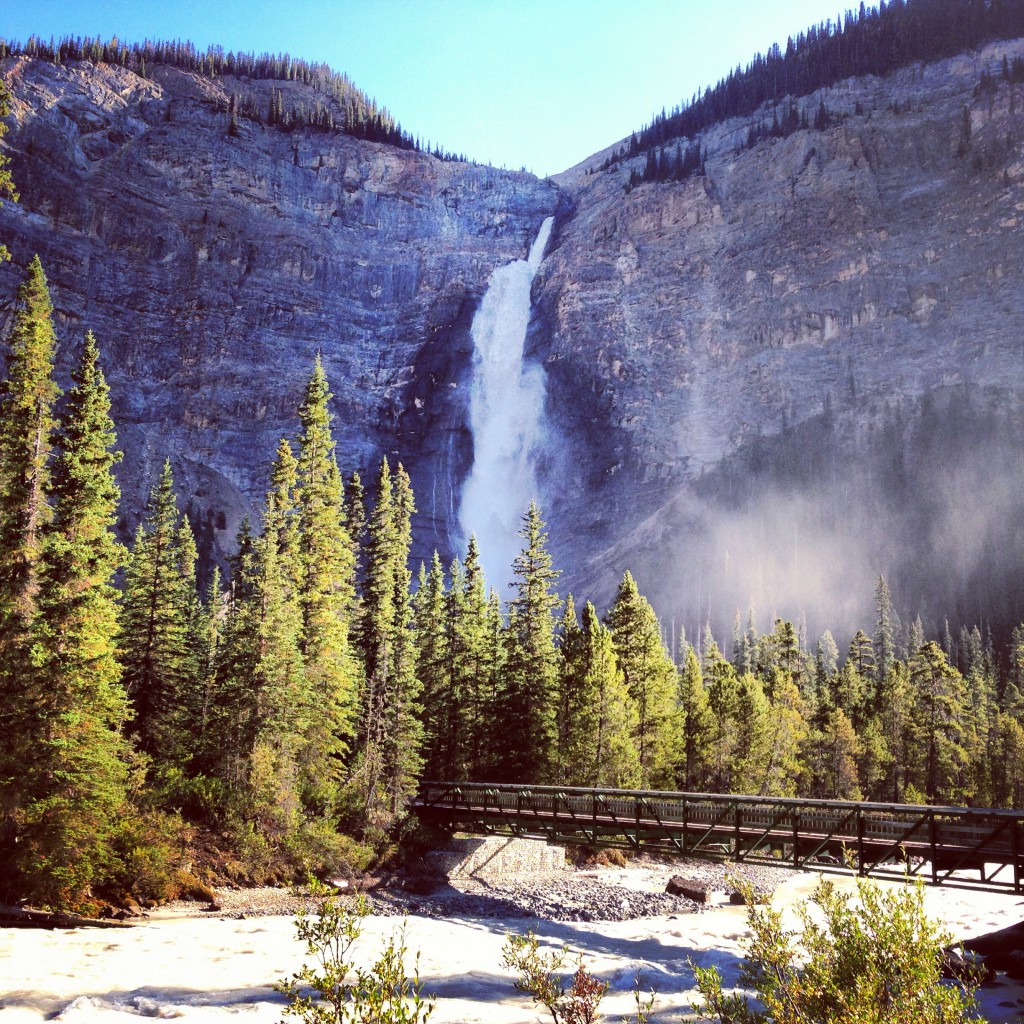 A bridge leads through a dense forest towards a waterfall cascading down a rocky mountain.