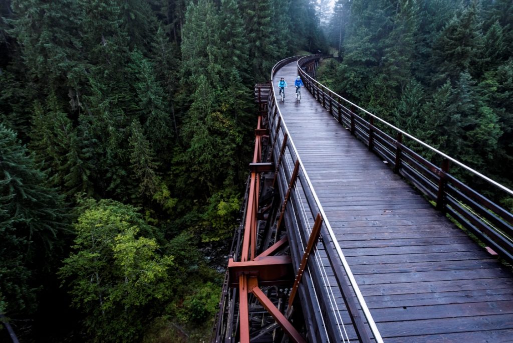 Two cyclists pedal across a trestle bridge through the forest on an overcast day. They are dressed in blue raincoats. 