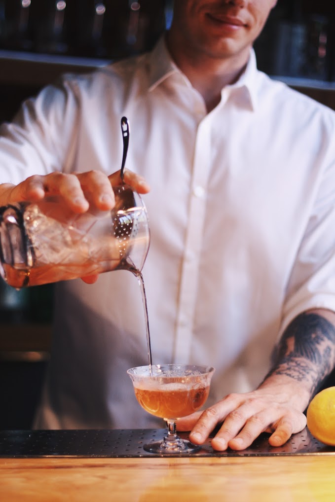 A bartender pours a cocktail from a shaker into a glass.