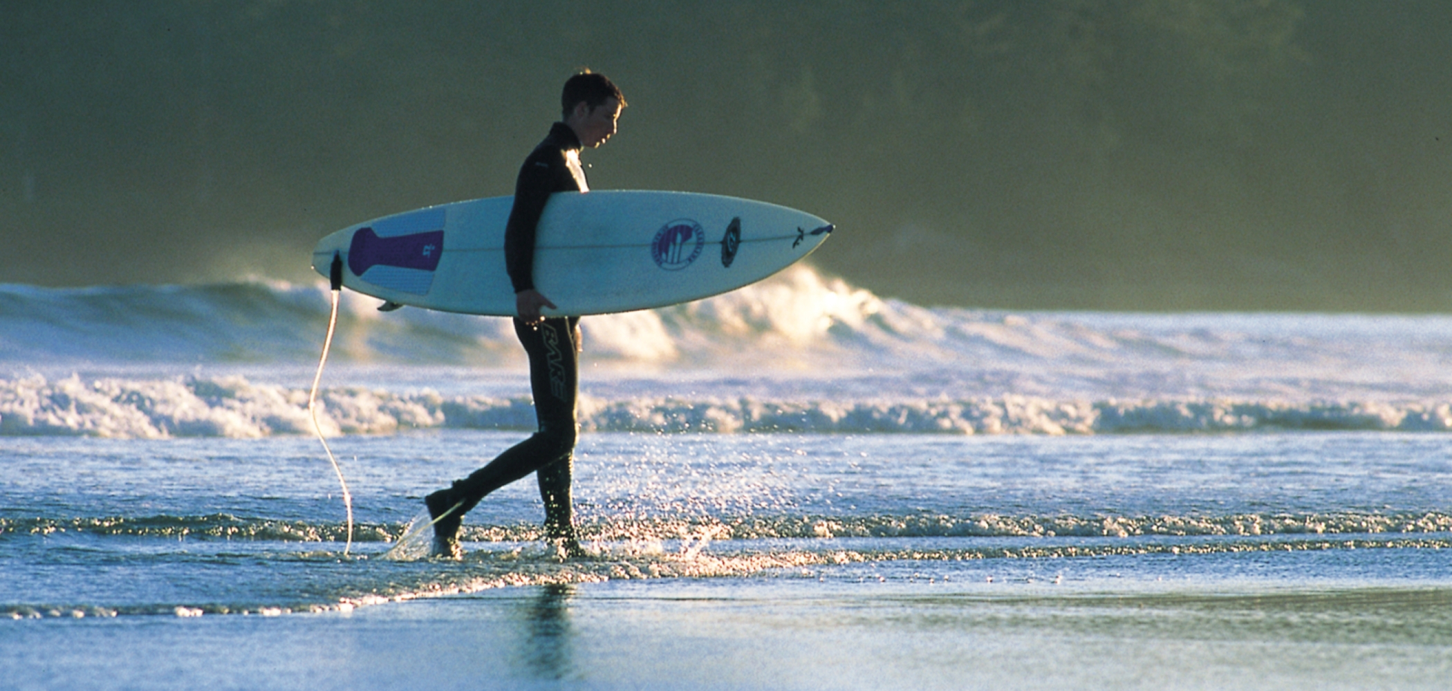 A man holds a surfboard under his arm as he walks along Long Beach in Tofino